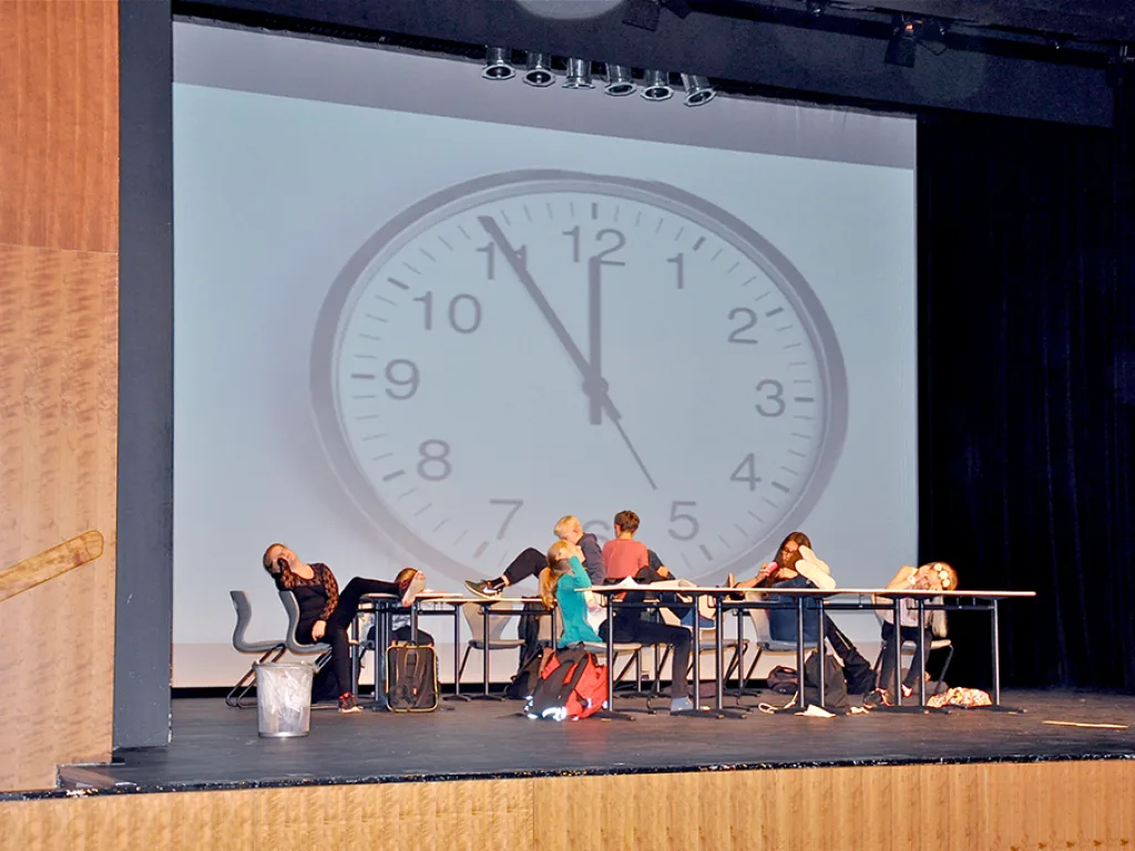 Eine Bühnenszene an Schultischen vor der Projektion einer großen Uhr.